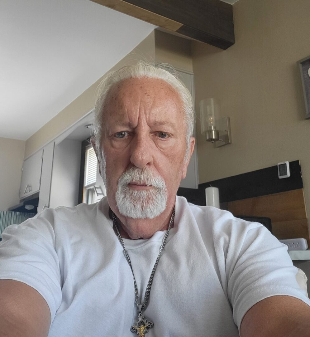 A man with white hair and beard sitting in front of a window.